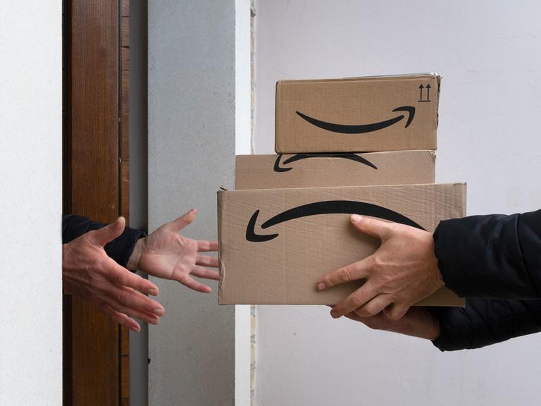 Drei Pakete mit dem Amazon Logo auf der Seite werden an einer Haustür überreicht.