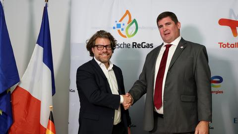 Zwei Männer reichen sich die Hand. Im Hintergrund sind die Logos der Unternehmen Deutsche ReGas und Total Energies zu sehen.