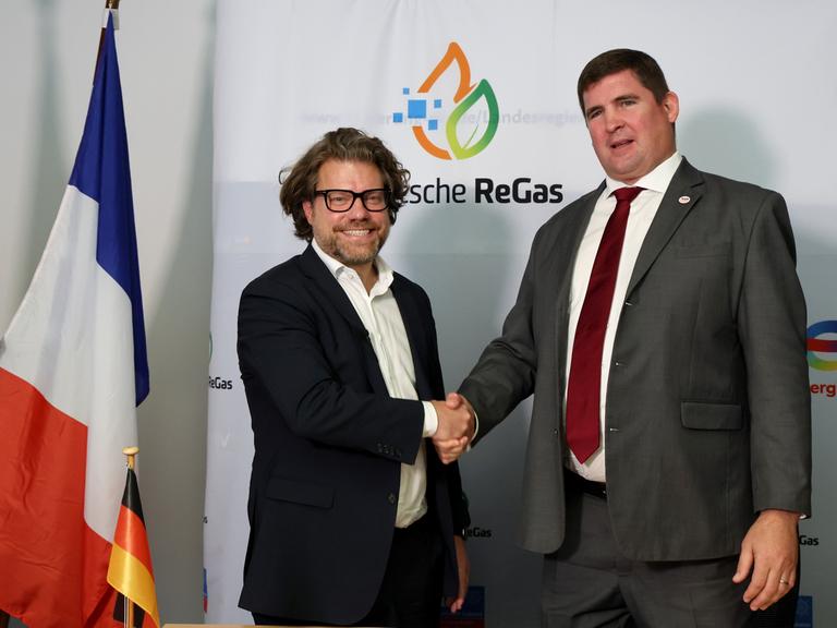 Zwei Männer reichen sich die Hand. Im Hintergrund sind die Logos der Unternehmen Deutsche ReGas und Total Energies zu sehen.