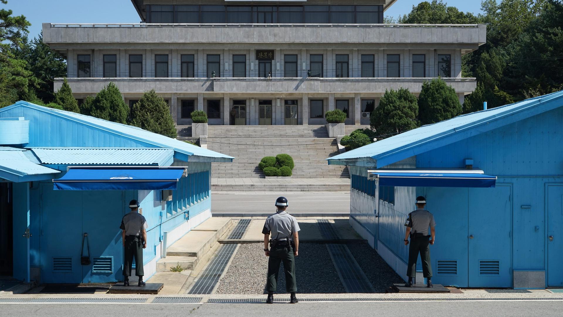 Militärische Siedlung 'Panmunjeom' in der demilitarisierten Zone (DMZ) zwischen Nord- und Südkorea. Rechts und links blaue Baracken, dazwischen verläuft die Grenze, im Vordergrund drei südkoreanische Grenzbeamte. Im Hintergrund ein Gebäude auf nordkoreanischer Seite.
