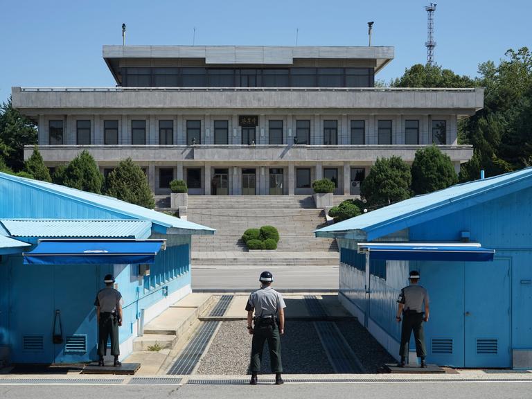 Militärische Siedlung 'Panmunjeom' in der demilitarisierten Zone (DMZ) zwischen Nord- und Südkorea. Rechts und links blaue Baracken, dazwischen verläuft die Grenze, im Vordergrund drei südkoreanische Grenzbeamte. Im Hintergrund ein Gebäude auf nordkoreanischer Seite.