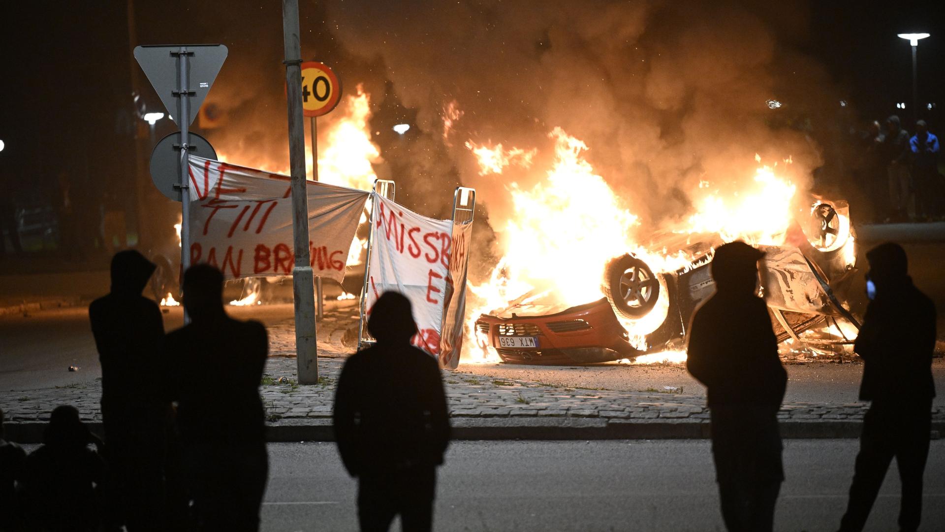 Schweden, Malmö; Unruhen nach Koranverbrennung. Ein Auto steht Kopf, es brennt. Menschen stehen davor.
