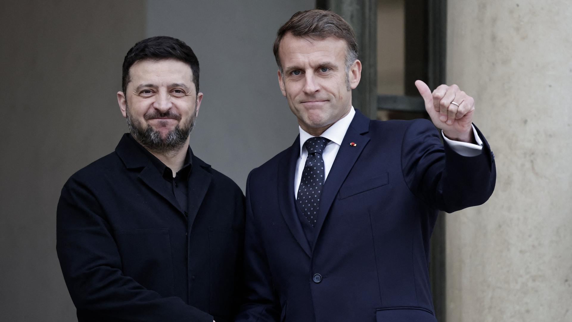 Der französische Präsident Emmanuel Macron (R) begrüßt seinen ukrainischen Amtskollegen Wolodymyr Selenskyj. Beide lächeln und schauen in die Kamera. 