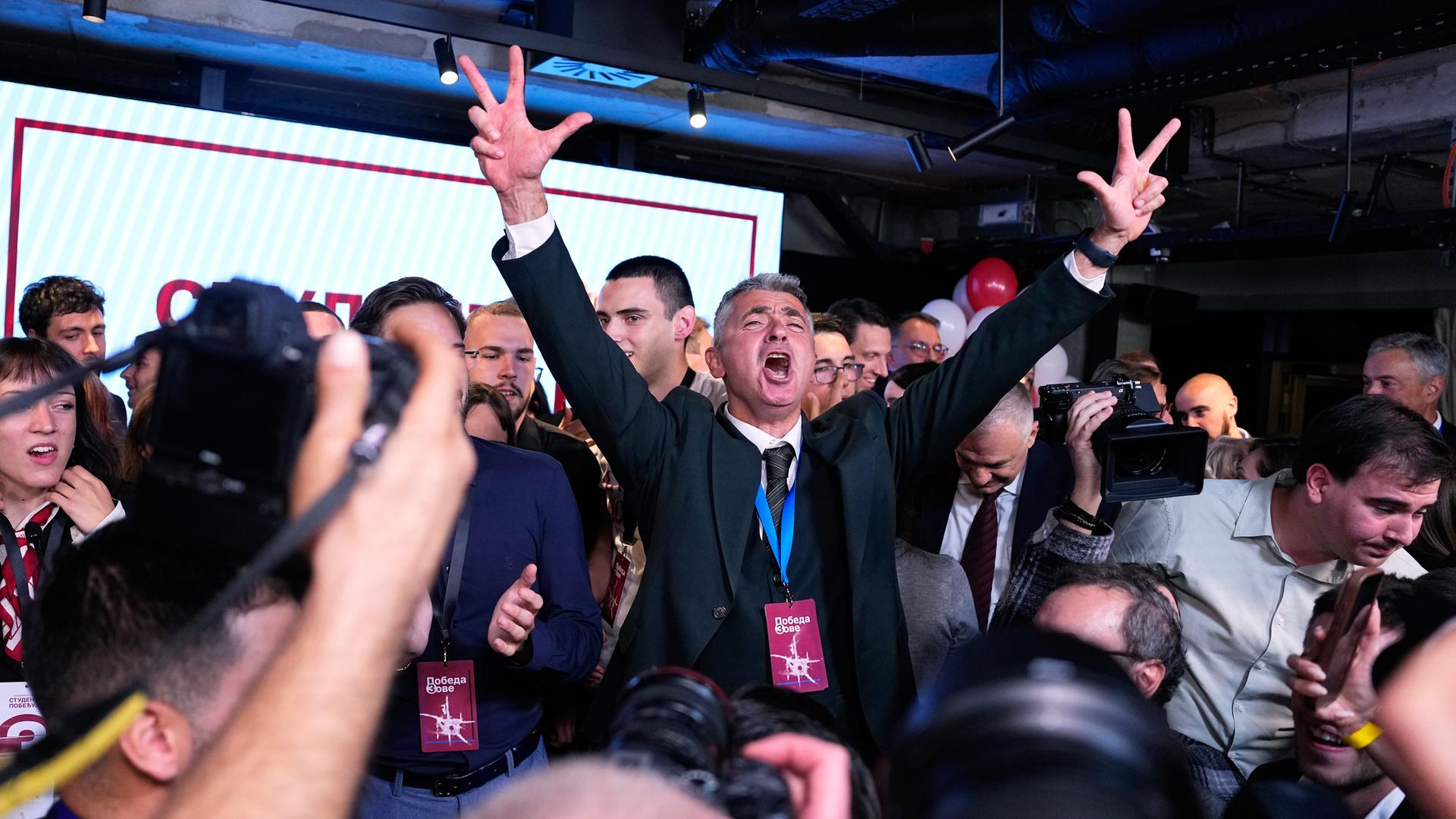 Ilija Srdanovic steht nach der Wahl zwischen Anhängern und Journalisten und feiert den Sieg, er zeigt mit beiden Händen den Drei-Finger-Gruß.