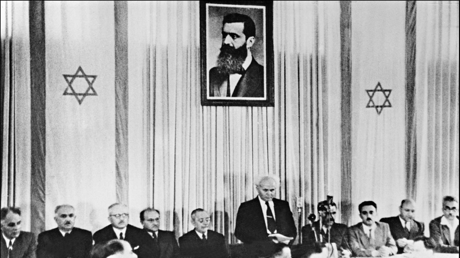 Der erste israelische Premierminister David Ben-Gurion (stehend) verkündet am 14. Mai 1948 in Tel Aviv vor Mitgliedern der jüdischen Ratsversammlung die Gründung des Staates Israel; oben ein Porträt von Theodor Herzl, Begründer des politischen Zionismus.