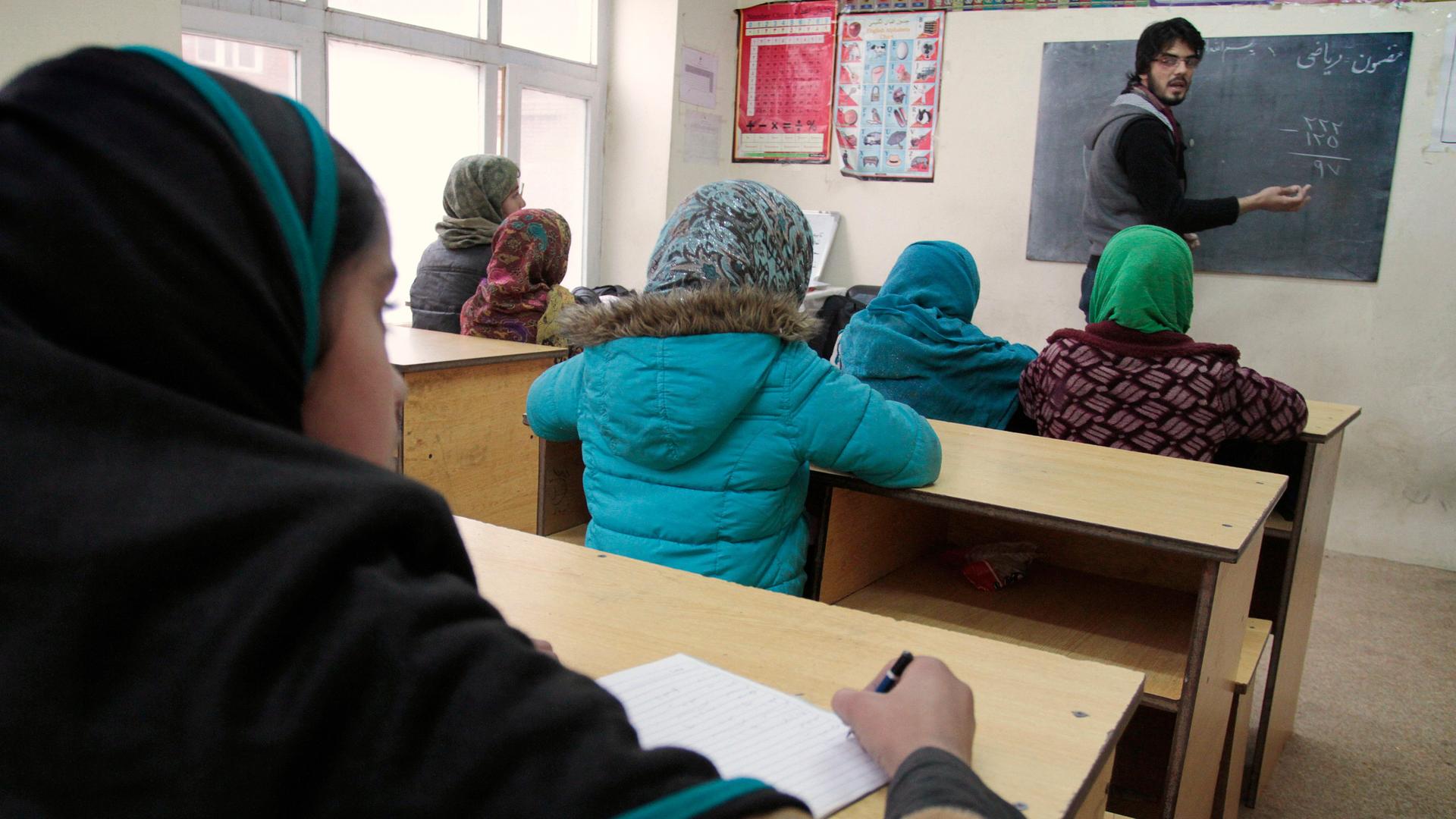 Ein Lehrer schreibt etwas an eine Tafel in einem Klassenzimmer in Afghanistan. Sechs Mädchen mit Kopftüchern sitzen auf ihren Plätzen.