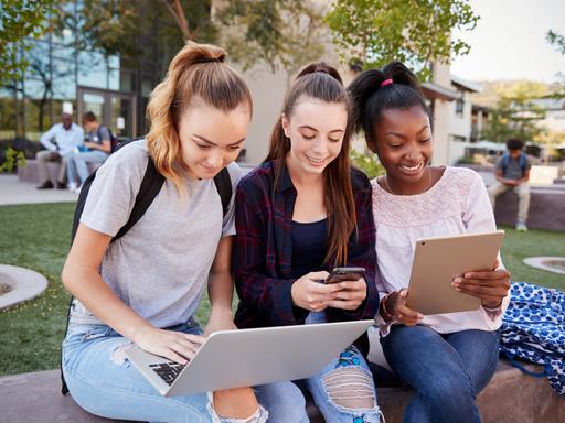 Drei Mädchen sitzen an einer Grünfläche nebeneinander, jede hat ein anderes Gerät in der Hand: Laptop, Smartphone, Tablet.