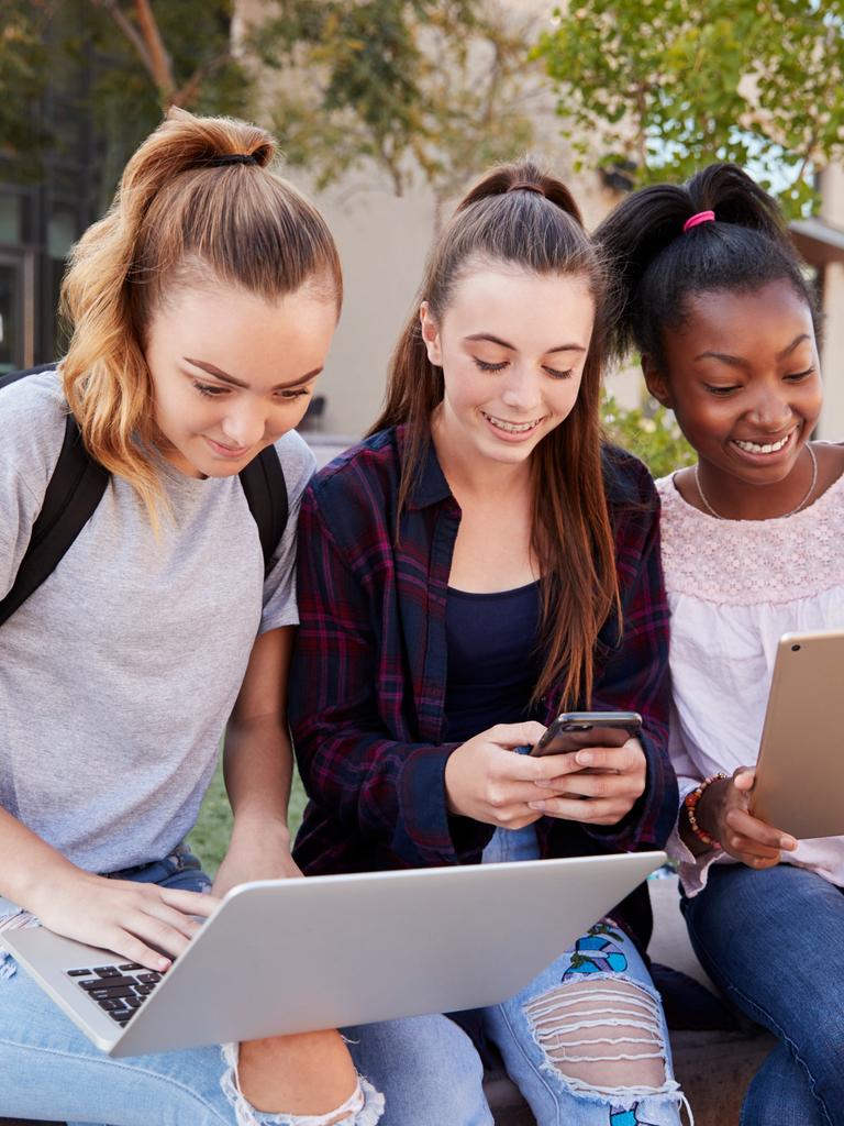 Drei Mädchen sitzen an einer Grünfläche nebeneinander, jede hat ein anderes Gerät in der Hand: Laptop, Smartphone, Tablet.