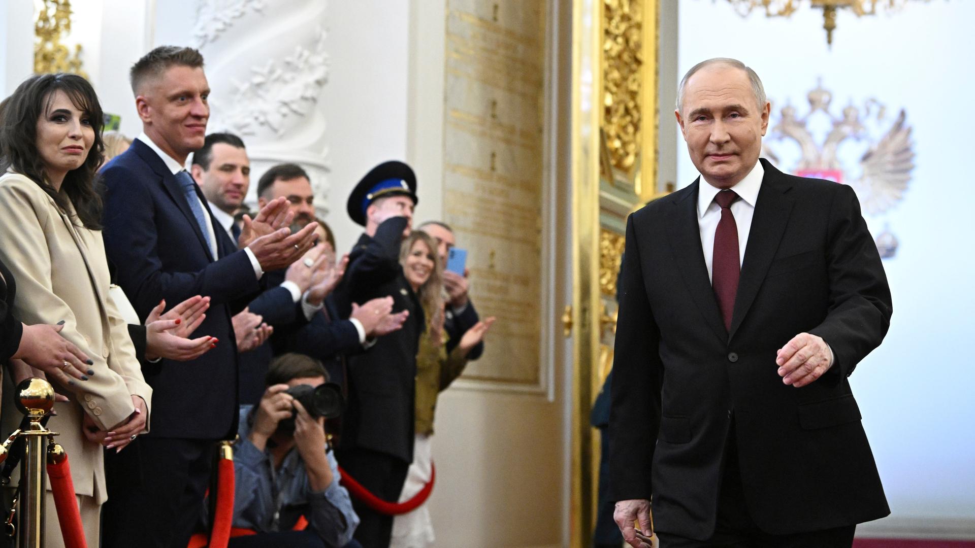 Wladimir Putin läuft durch eine prunkvolle Halle. Zuschauer bejubeln ihn.