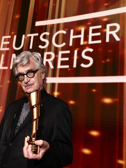 Wim Wender hält auf der Bühne den Ehrenpreis des Deutschen Filmpreises in der Hand. Hinter ihm steht in großer weißer Schrift "Deutscher Filmpreis".
