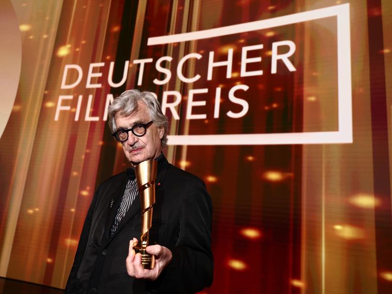 Wim Wender hält auf der Bühne den Ehrenpreis des Deutschen Filmpreises in der Hand. Hinter ihm steht in großer weißer Schrift "Deutscher Filmpreis".
