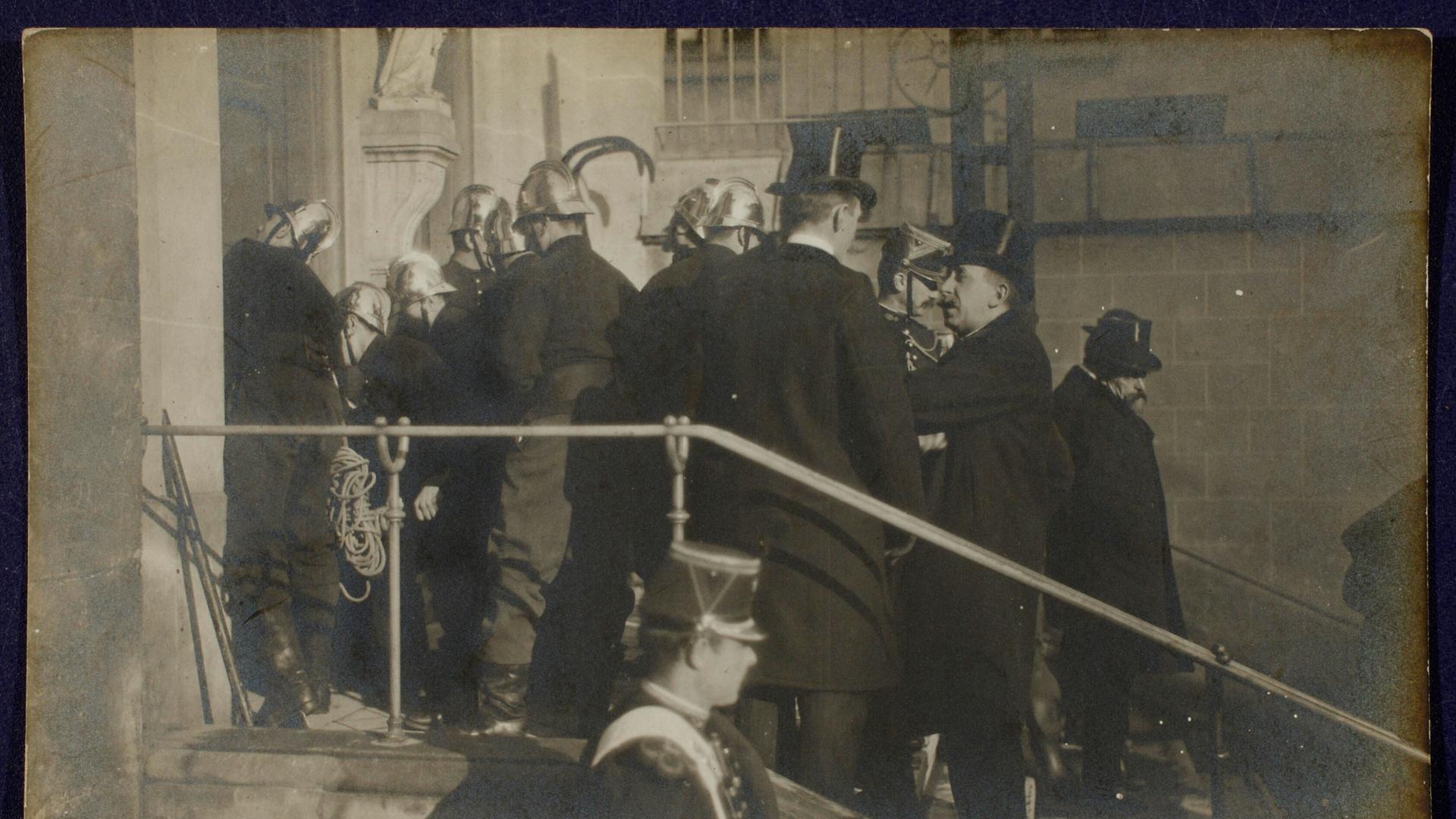 Szene auf einer Fotopostkarte: Polizisten und Feuerwehrleute in Uniformen und Helmen stehen auf einer Treppe vor einer Kirche. Sie sind dabei, die Türen aufzubrechen. Szene auf einer Fotopostkarte: Polizisten und Feuerwehrleute in Uniformen und Helmen stehen auf einer Treppe vor einer Kirche. Sie sind dabei, die Türen aufzubrechen.