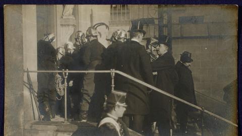 Szene auf einer Fotopostkarte: Polizisten und Feuerwehrleute in Uniformen und Helmen stehen auf einer Treppe vor einer Kirche. Sie sind dabei, die Türen aufzubrechen.