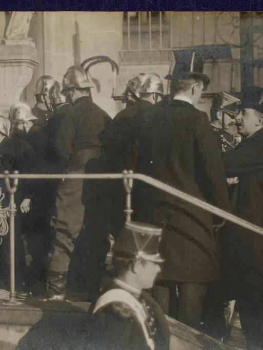 Szene auf einer Fotopostkarte: Polizisten und Feuerwehrleute in Uniformen und Helmen stehen auf einer Treppe vor einer Kirche. Sie sind dabei, die Türen aufzubrechen. Szene auf einer Fotopostkarte: Polizisten und Feuerwehrleute in Uniformen und Helmen stehen auf einer Treppe vor einer Kirche. Sie sind dabei, die Türen aufzubrechen.