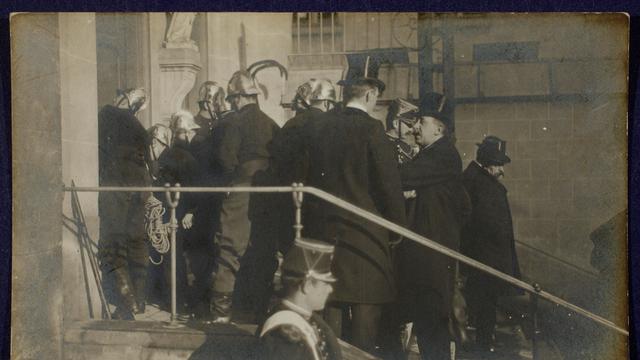 Szene auf einer Fotopostkarte: Polizisten und Feuerwehrleute in Uniformen und Helmen stehen auf einer Treppe vor einer Kirche. Sie sind dabei, die Türen aufzubrechen. Szene auf einer Fotopostkarte: Polizisten und Feuerwehrleute in Uniformen und Helmen stehen auf einer Treppe vor einer Kirche. Sie sind dabei, die Türen aufzubrechen.