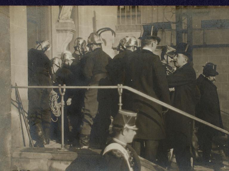 Szene auf einer Fotopostkarte: Polizisten und Feuerwehrleute in Uniformen und Helmen stehen auf einer Treppe vor einer Kirche. Sie sind dabei, die Türen aufzubrechen. Szene auf einer Fotopostkarte: Polizisten und Feuerwehrleute in Uniformen und Helmen stehen auf einer Treppe vor einer Kirche. Sie sind dabei, die Türen aufzubrechen.