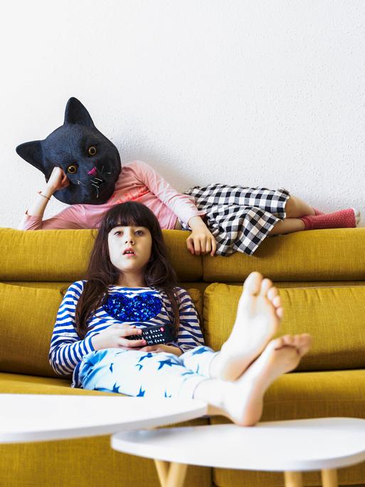 Zwei gelangweilte Mädchen sitzen auf der Couch und sehen fern, eine trägt eine Katzenmaske.