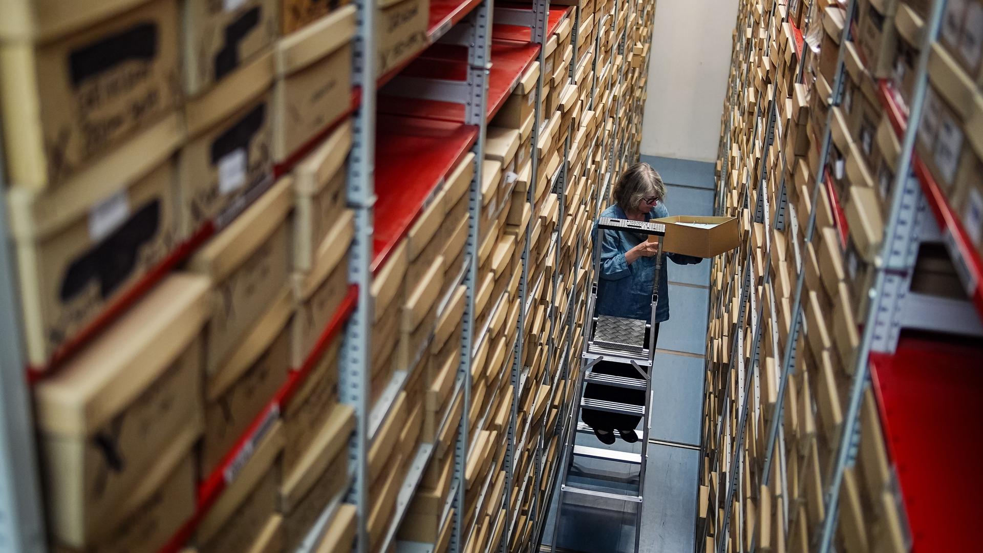 Eine Frau steht auf einer Leiter und nimmt Akten aus einem Archivregal. 