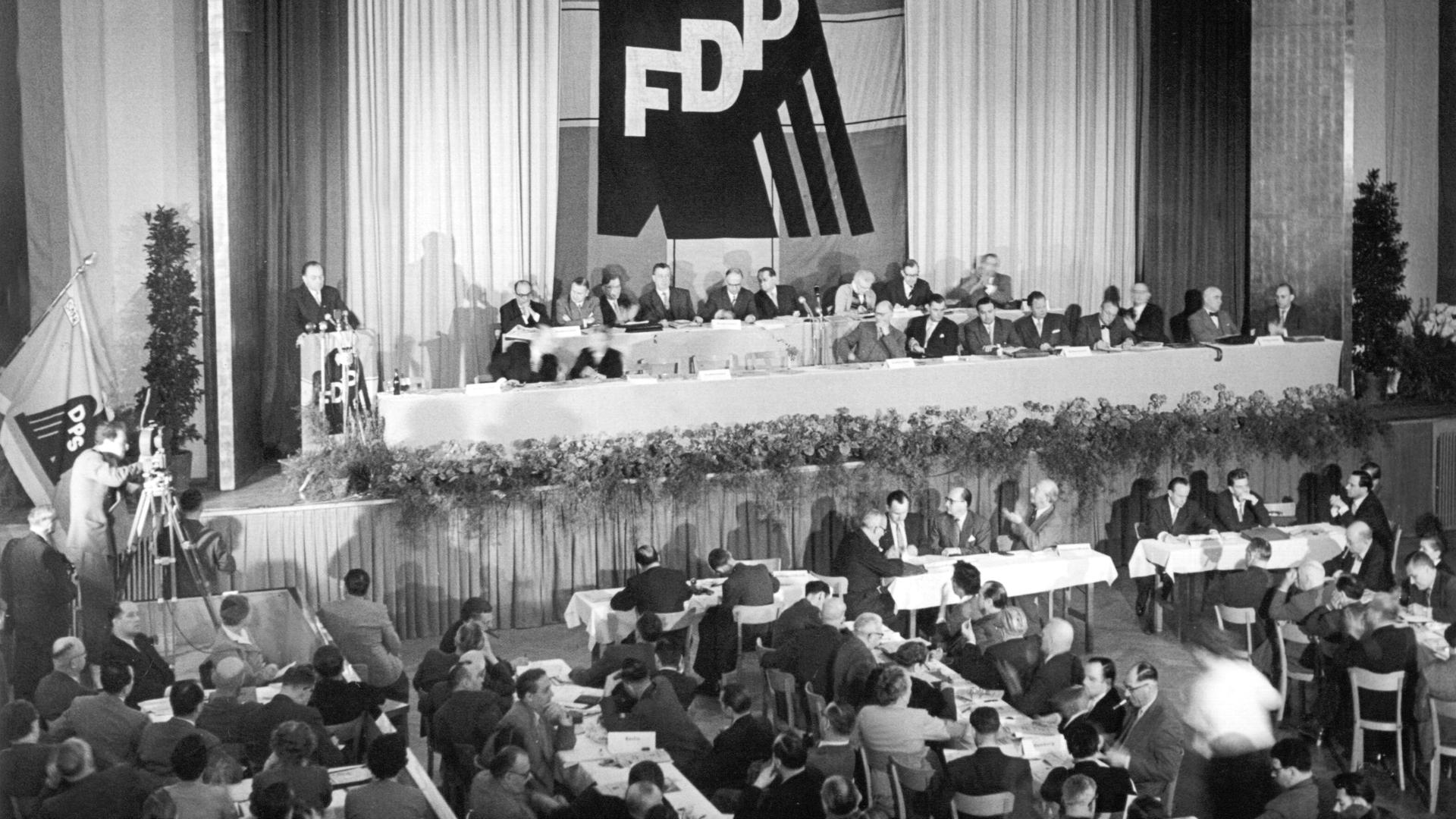 Schwarz-Weiß-Aufnahme vom Parteitag der FDP. Mehrere Menschen, überwiegend Männer, sitzen in einem Saal. Über der Bühne hängt das Logo der FDP. Schwarz-Weiß-Aufnahme vom Parteitag der FDP. Mehrere Menschen, überwiegend Männer, sitzen in einem Saal. Über der Bühne hängt das Logo der FDP.