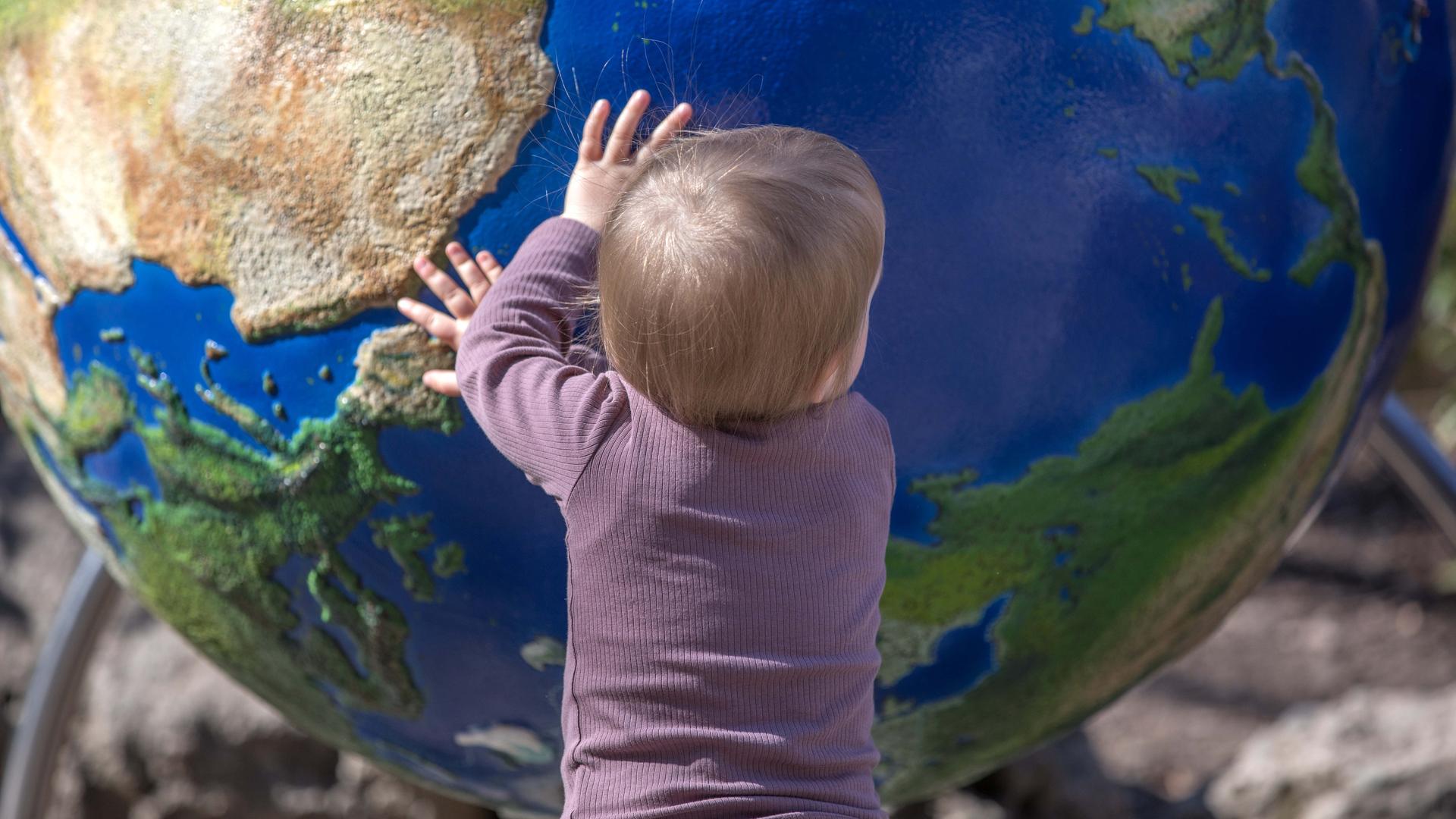 Ein kleines Kind steht vor einer Weltkugel und bewegt den Globus mit den Händen.