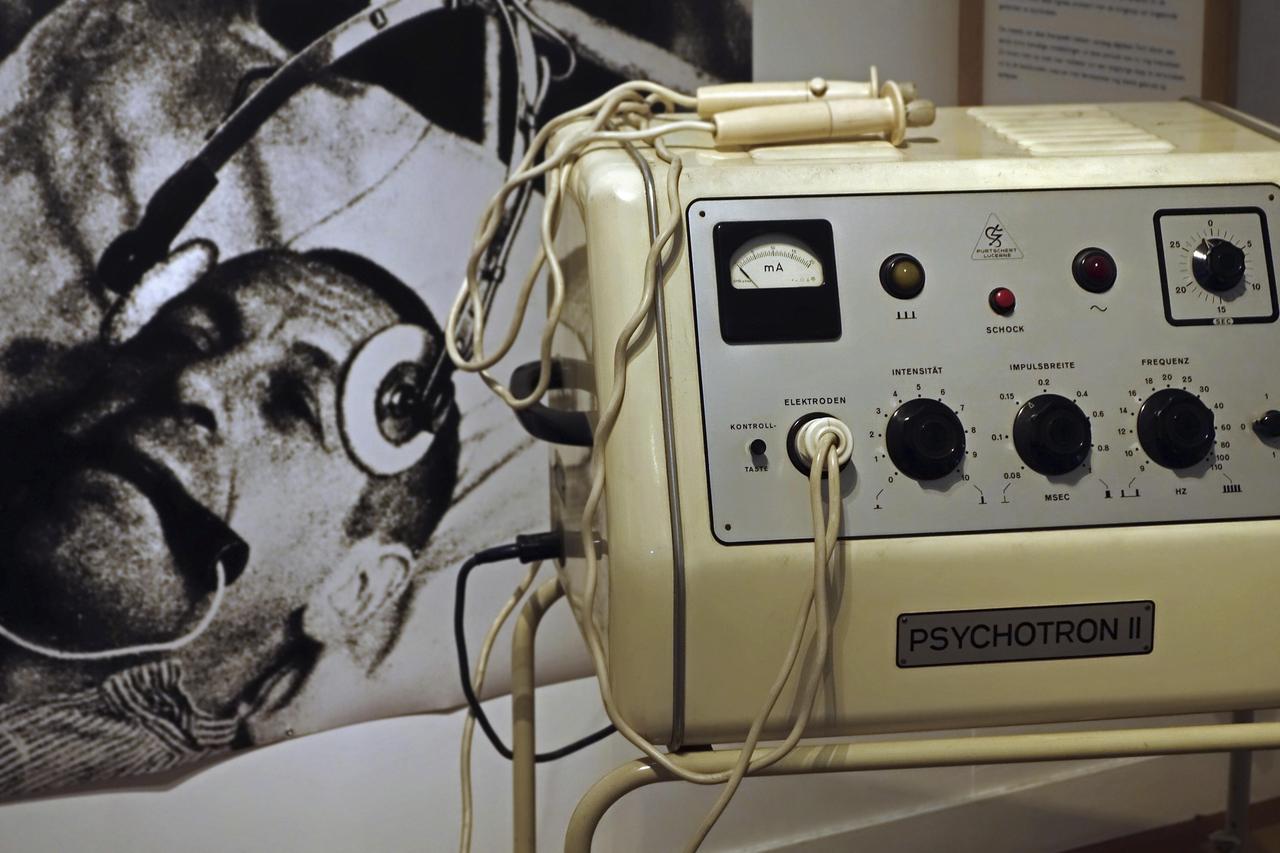 Ein historisches medizinisches Messgerät mit der Aufschrift "Psychotron II". Dahinter an der Wand hängt ein Foto, auf dem ein Mann Elektroden am Kopf und eine Art Beißgummi im Mund hat. Ein historisches medizinisches Messgerät mit der Aufschrift "Psychotron II". Dahinter an der Wand hängt ein Foto, auf dem ein Mann Elektroden am Kopf und eine Art Beißgummi im Mund hat.