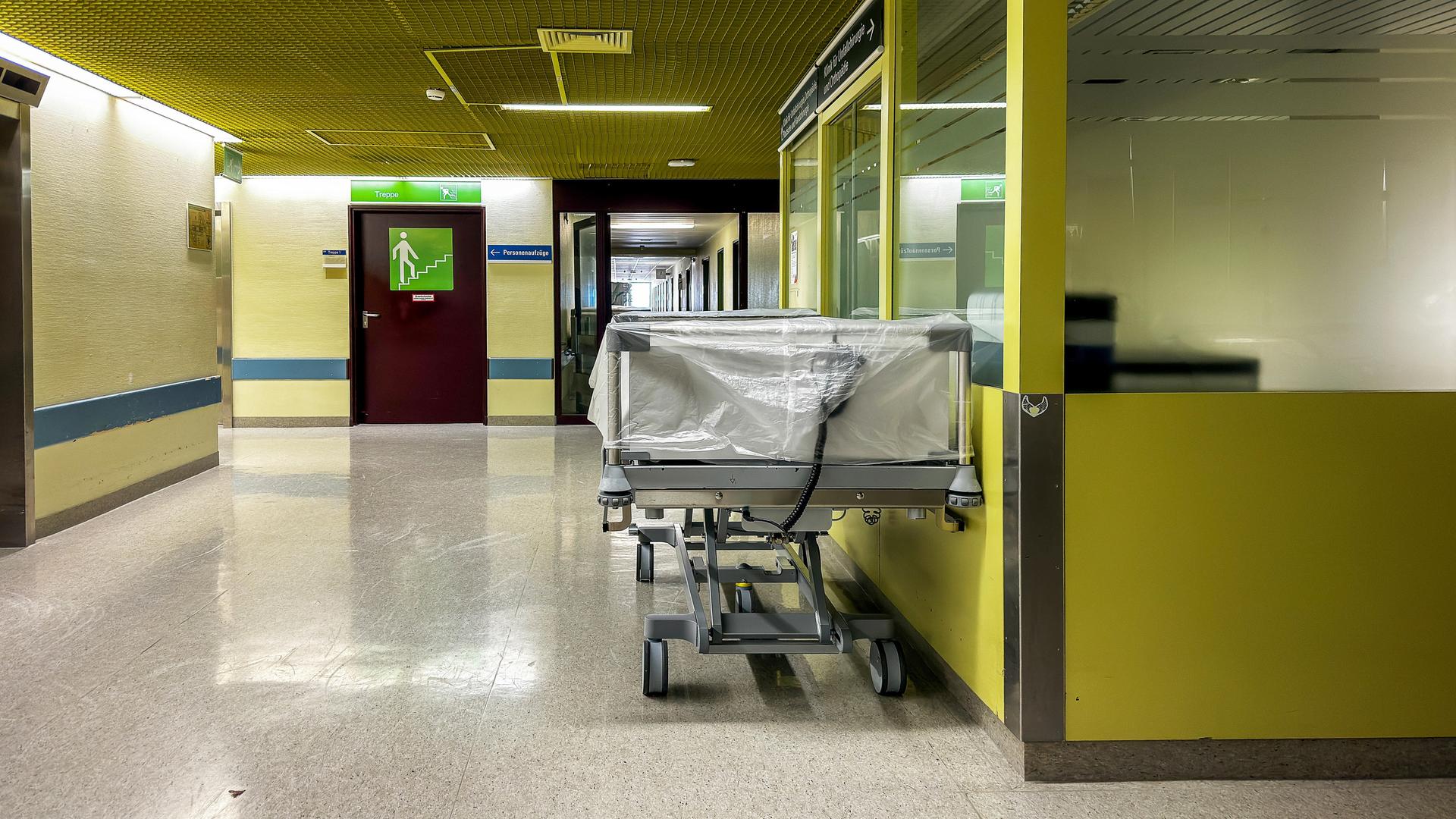Ein leeres Krankenhausbett steht auf einem mit Neonlicht beleuchteten gelben Klinikflur. Ein leeres Krankenhausbett steht auf einem mit Neonlicht beleuchteten gelben Klinikflur.