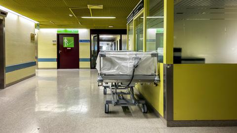 Ein leeres Krankenhausbett steht auf einem mit Neonlicht beleuchteten gelben Klinikflur.