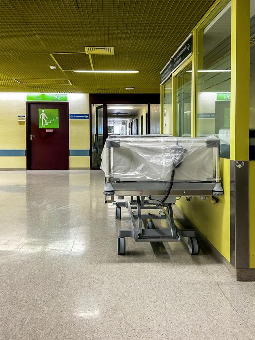 Ein leeres Krankenhausbett steht auf einem mit Neonlicht beleuchteten gelben Klinikflur.
