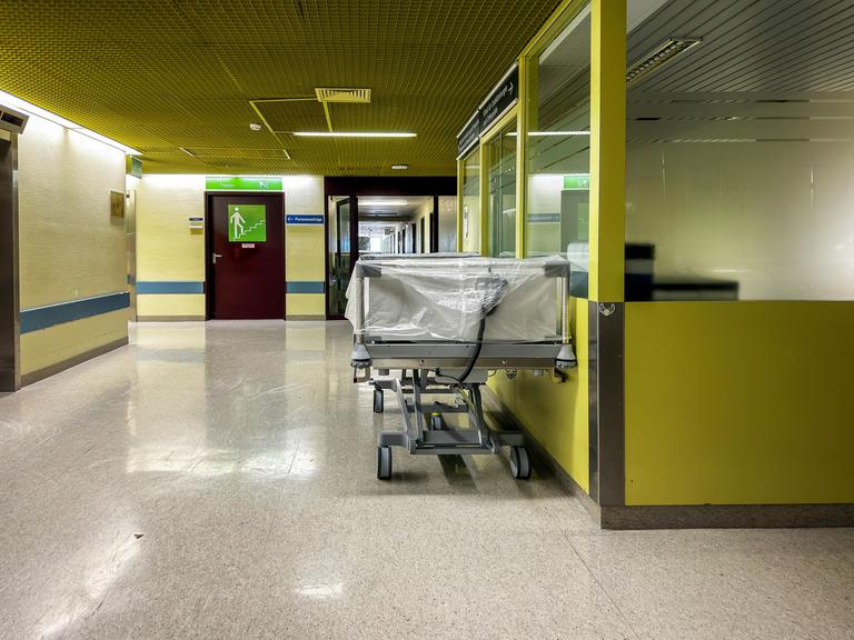 Ein leeres Krankenhausbett steht auf einem mit Neonlicht beleuchteten gelben Klinikflur.