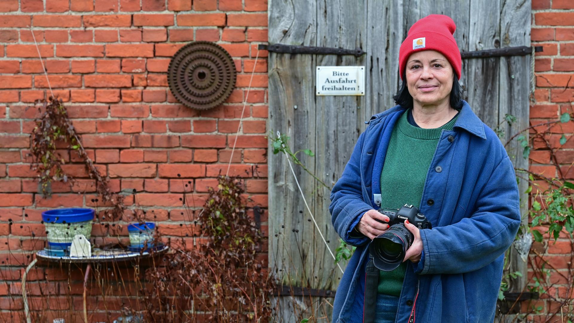 Die Fotografin Isabel Kittler steht mit einer Kamera vor einer Scheune in ihrem Garten in der Uckermark. Sie trägt eine rote Mütze und eine blaue Jacke. 