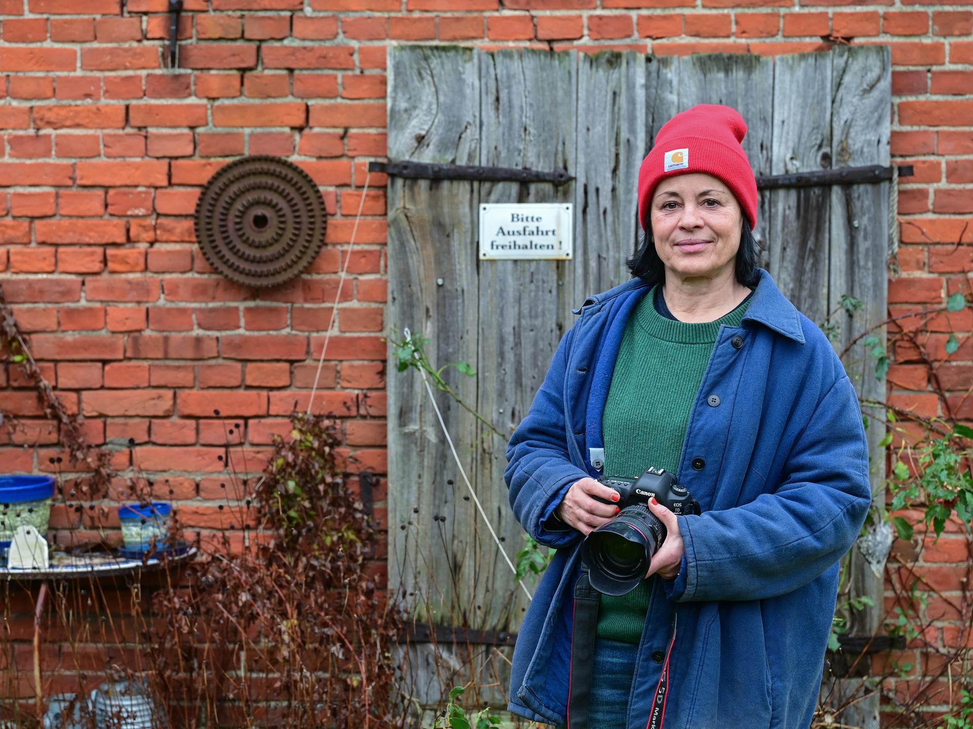 Die Fotografin Isabel Kittler steht mit einer Kamera vor einer Scheune in ihrem Garten in der Uckermark. Sie trägt eine rote Mütze und eine blaue Jacke. Die Fotografin Isabel Kittler steht mit einer Kamera vor einer Scheune in ihrem Garten in der Uckermark. Sie trägt eine rote Mütze und eine blaue Jacke.
