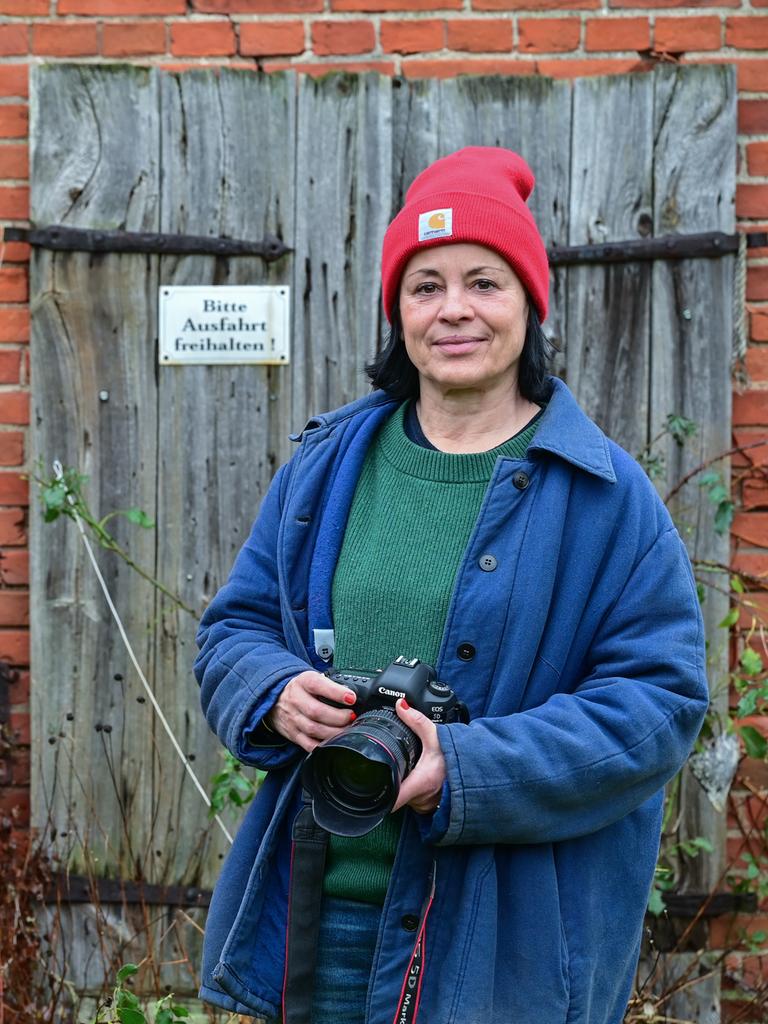 Die Fotografin Isabel Kittler steht mit einer Kamera vor einer Scheune in ihrem Garten in der Uckermark. Sie trägt eine rote Mütze und eine blaue Jacke. Die Fotografin Isabel Kittler steht mit einer Kamera vor einer Scheune in ihrem Garten in der Uckermark. Sie trägt eine rote Mütze und eine blaue Jacke.