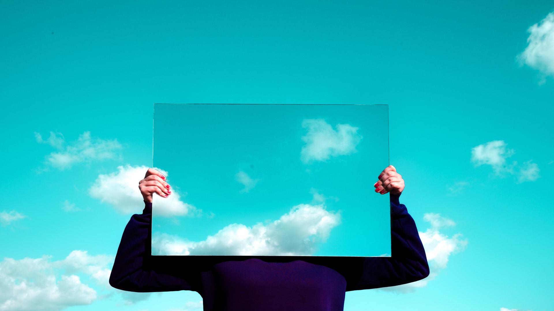 Eine Frau hält einen rechteckigen Spiegel vor ihr Gesicht, in dem sich die Wolken spiegeln. 