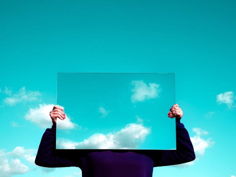 Eine Frau hält einen rechteckigen Spiegel vor ihr Gesicht, in dem sich die Wolken spiegeln. 