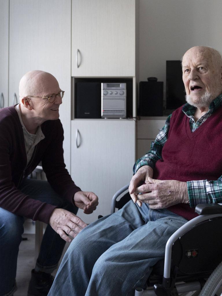 Ein alter Mann sitzt im Pflegeheim im Rollstuhl, ein anderer Mann sitzt neben ihm. Beide sind im Gespräch. 