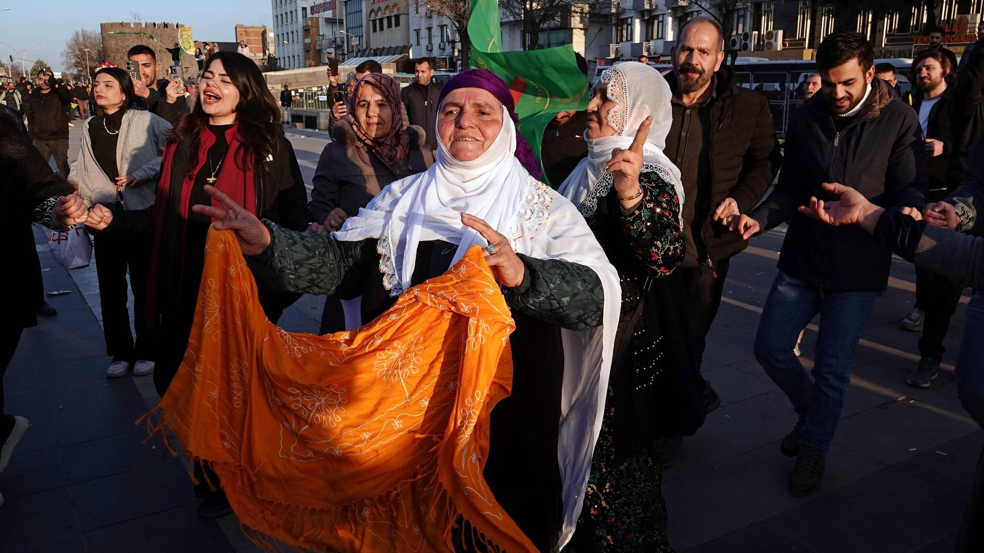 Eine Straßenszene in Diyarbakir in der Türkei: Eine ältere mutmaßliche Kurdin mit weißem Kopftuch und einem orange Tuch in den Händen tanzt neben anderen ähnlich  gekleideten Frauen und blickt erwartungsvoll nach vorn. 