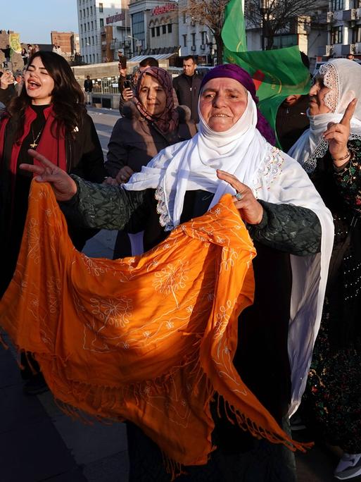 Eine Straßenszene in Diyarbakir in der Türkei: Eine ältere mutmaßliche Kurdin mit weißem Kopftuch und einem orange Tuch in den Händen tanzt neben anderen ähnlich  gekleideten Frauen und blickt erwartungsvoll nach vorn. 