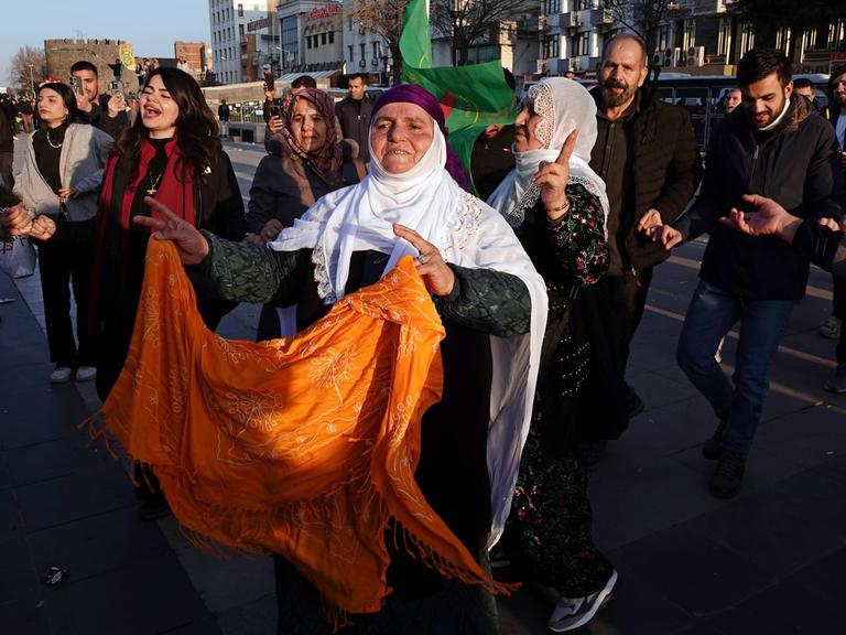 Eine Straßenszene in Diyarbakir in der Türkei: Eine ältere mutmaßliche Kurdin mit weißem Kopftuch und einem orange Tuch in den Händen tanzt neben anderen ähnlich  gekleideten Frauen und blickt erwartungsvoll nach vorn. 