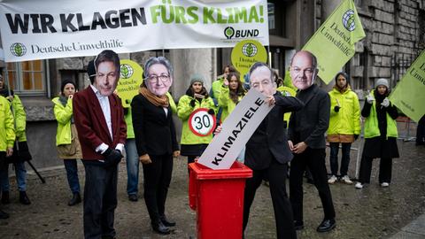Ein Aktivist mit einer Maske von Verkehrsminister Wissing (M) wirft neben Aktivisten mit Masken von Wirtschaftsminister Habeck (l-r), Bauministerin Geywitz, und Bundeskanzler Scholz vor einer Verhandlung vor dem Oberverwaltungsgericht Berlin über eine Klage der Deutschen Umwelthilfe (DUH) und des Bunds für Umwelt und Naturschutz Deutschland (BUND) gegen die Bundesregierung ein Schild mit der Aufschrift "Klimaziele" in eine Tonne. 