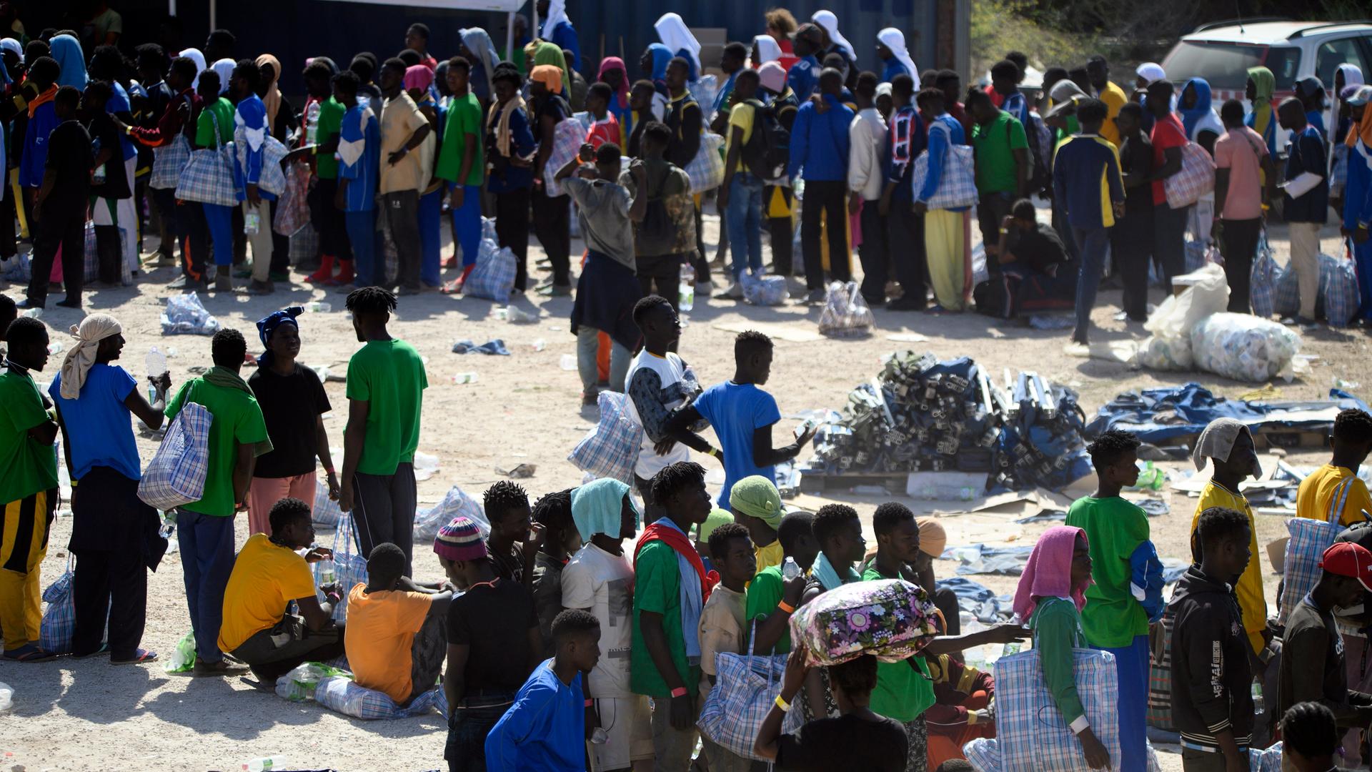 Migranten mit Kleiderbündeln und Taschen stehen in langen Schlangen auf staubigem Boden im Aufnahmezentrum auf der italienischen Insel Lampedusa.