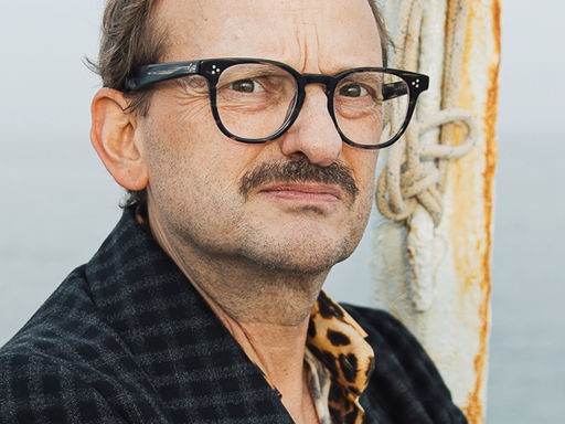 Milan Peschel trägt ein kariertes Sacko und schaut streng in die Kamera. Im Hintergrund ist das offene Meer zu sehen.