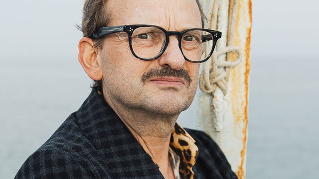 Milan Peschel trägt ein kariertes Sacko und schaut streng in die Kamera. Im Hintergrund ist das offene Meer zu sehen.