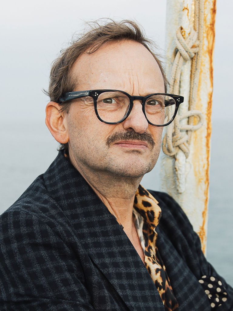 Milan Peschel trägt ein kariertes Sacko und schaut streng in die Kamera. Im Hintergrund ist das offene Meer zu sehen.