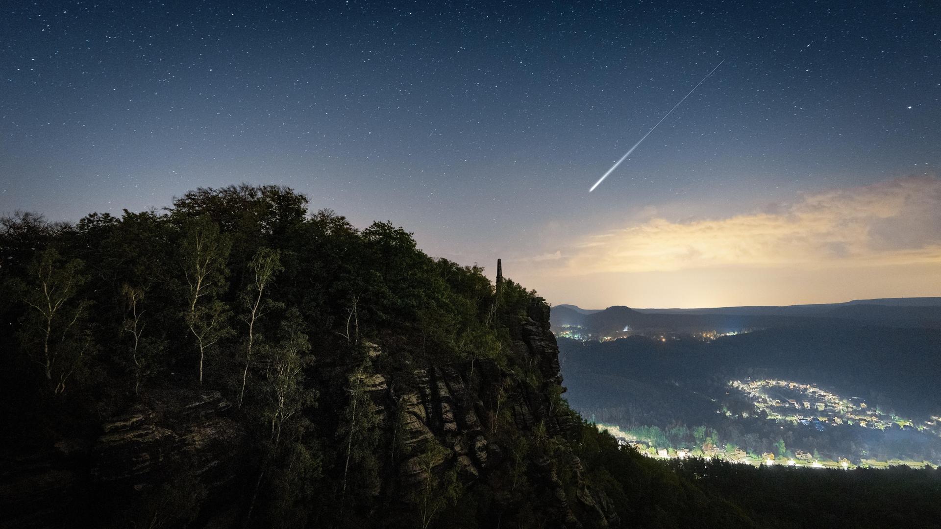 Im Vordergrund die schwarze Silhouette eines bewaldeten Berghangs, in der Ferne die Lichter eines Ortes in einem Tal. Darüber ein Nachthimmel mit unzähligen Sternen, durch den sich der helle Strich der Sternschnuppe zieht. Im Vordergrund die schwarze Silhouette eines bewaldeten Berghangs, in der Ferne die Lichter eines Ortes in einem Tal. Darüber ein Nachthimmel mit unzähligen Sternen, durch den sich der helle Strich der Sternschnuppe zieht.