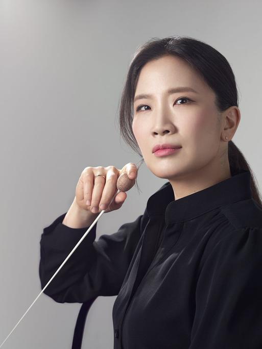 Eun Sun Kim blickt ernst in die Kamera und hält einen Dirigentenstab in der Hand.