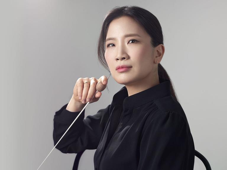 Eun Sun Kim blickt ernst in die Kamera und hält einen Dirigentenstab in der Hand.