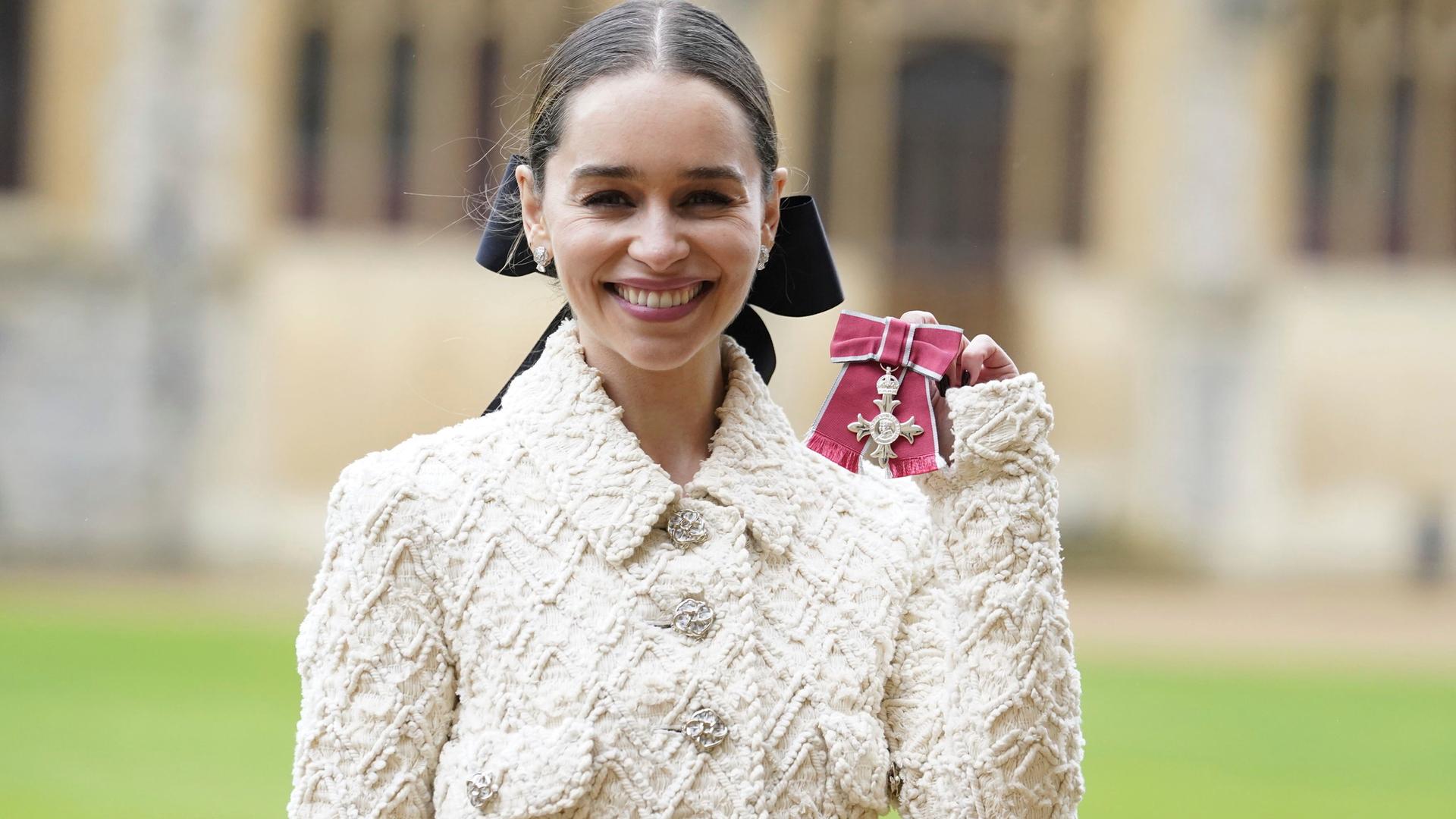Emilia Clark lächelt und hält ihren Orden des Britischen Empire in die Höhe.