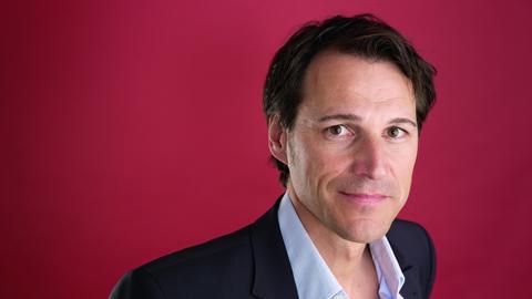 Ein Mann mit dunkelbraunem Haar und dunkelbraunen steht vor einem dunkelroten Hintergrund und blickt freundlich in die Kamera. Er trägt ein hellblaues Hemd und ein schwarzes Jackett darüber. 