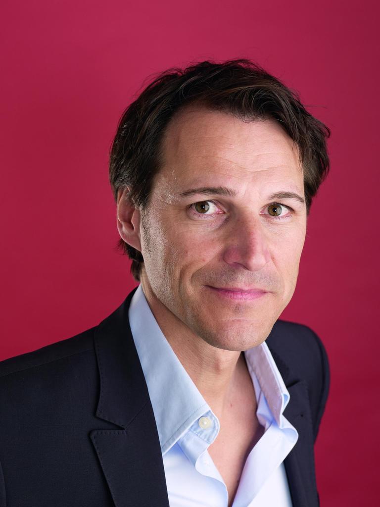 Ein Mann mit dunkelbraunem Haar und dunkelbraunen steht vor einem dunkelroten Hintergrund und blickt freundlich in die Kamera. Er trägt ein hellblaues Hemd und ein schwarzes Jackett darüber. 