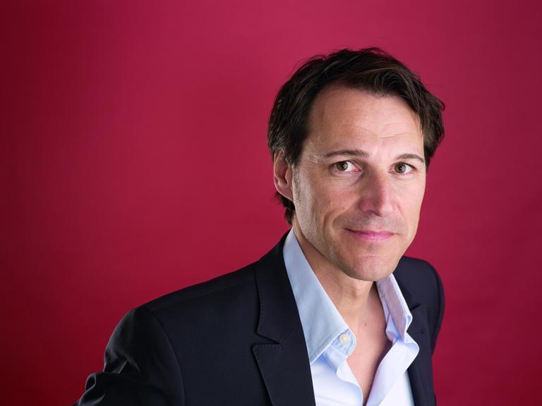 Ein Mann mit dunkelbraunem Haar und dunkelbraunen steht vor einem dunkelroten Hintergrund und blickt freundlich in die Kamera. Er trägt ein hellblaues Hemd und ein schwarzes Jackett darüber. 